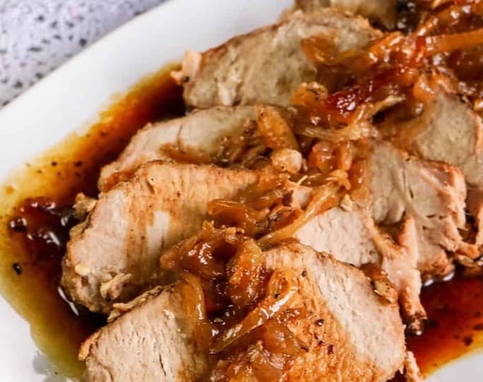 Sliced pork loin roast with caramelized onions and sauce on a white plate. A slow cooker is partially visible in the background, hinting at deliciously effortless crockpot recipes.