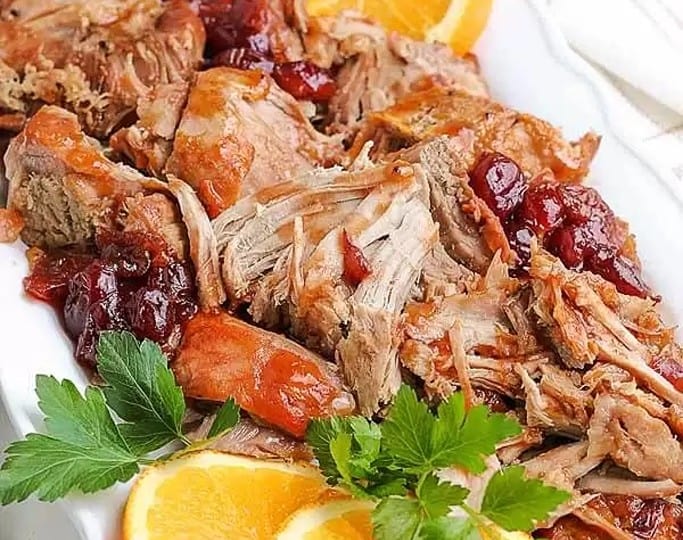 A plate of shredded pork loin, slow-cooked to perfection using a favorite crockpot recipe, is garnished with vibrant orange slices and parsley, then topped with tangy cranberries.