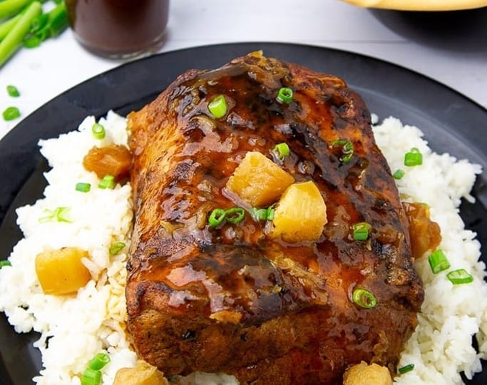 A glazed roast pork loin sits on white rice, topped with pineapple chunks and green onions. A pitcher of sauce and rolls are in the background, reminiscent of comforting crockpot recipes.