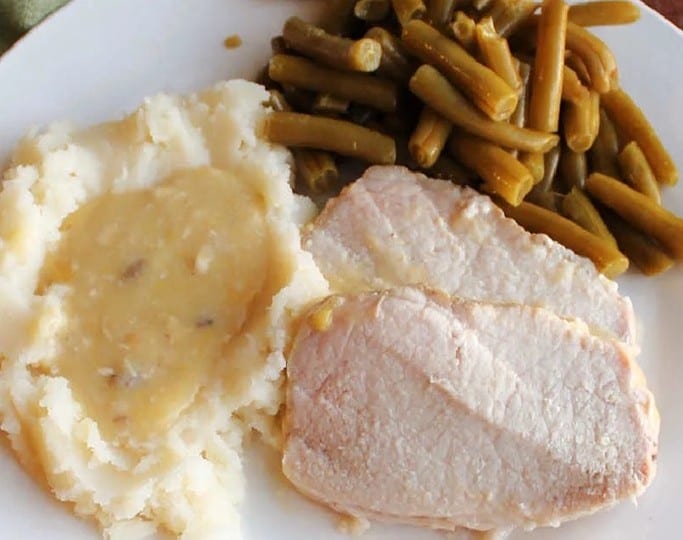 A white plate with sliced pork, mashed potatoes topped with gravy, and green beans showcases the richness of pork loin crockpot recipes. A green cloth is elegantly draped in the background.