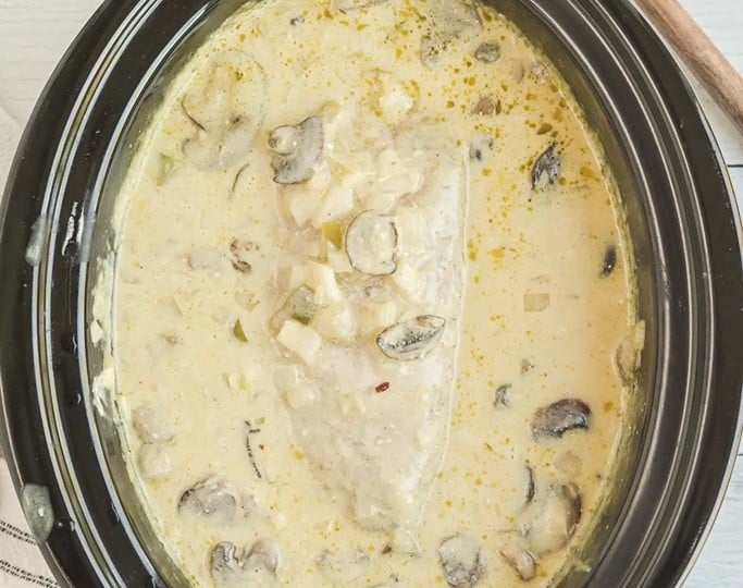 A creamy chicken and mushroom dish in a black slow cooker, reminiscent of pork loin crockpot recipes, sits enticingly next to a wooden spoon on a pristine white surface.