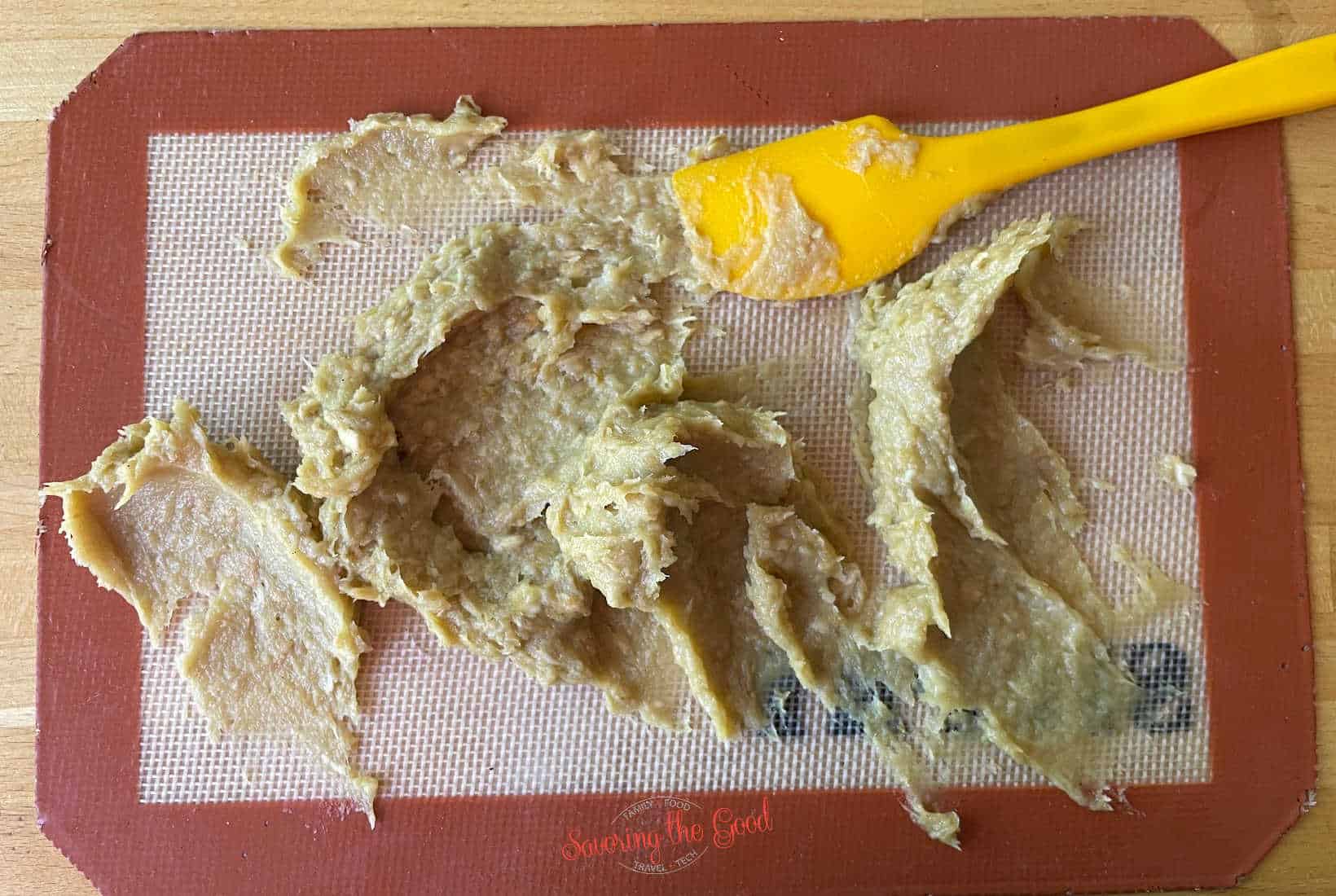 purred garlic paste being spread on a sil pat mat with a yellow spatula.