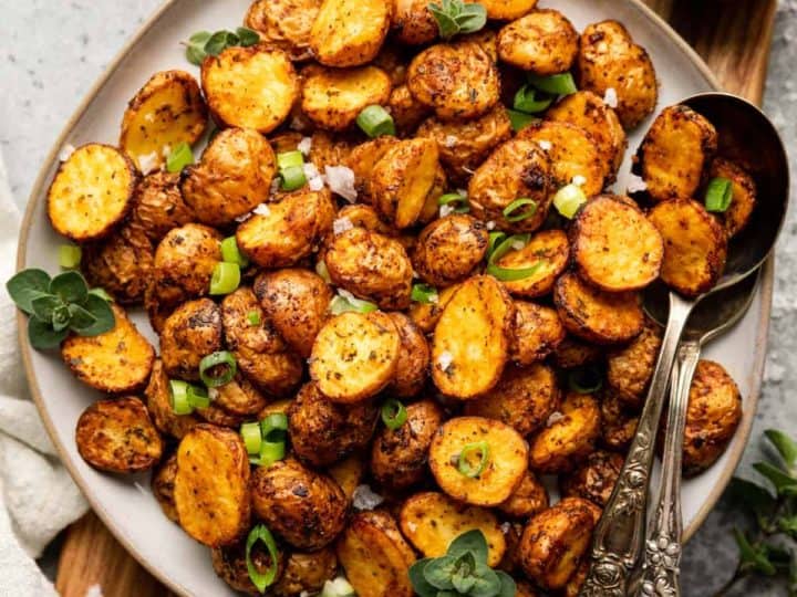 A plate of roasted baby potatoes garnished with green onions and herbs, with two serving spoons on the side.