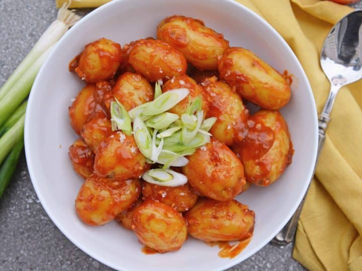 A bowl of baby potatoes in red sauce, garnished with sliced green onions, offers a delightful addition to your favorite recipes. A yellow napkin and spoon are neatly arranged beside the bowl.