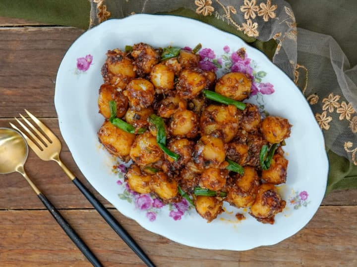 A plate of spicy roasted baby potatoes garnished with green onions, placed on a floral-patterned platter. Gold and black utensils are beside the dish on a wooden surface. Perfect for those seeking flavorful baby potatoes recipes.