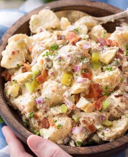A bowl of creamy potato salad features tender baby potatoes with diced bacon, red onion, celery, and herbs.