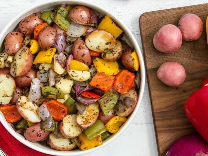 A bowl of roasted vegetables featuring baby potatoes, vibrant bell peppers, and onions. Nearby, sliced red and yellow peppers mingle with baby potatoes on a wooden board. A red cloth and spoon lie alongside this delightful scene.