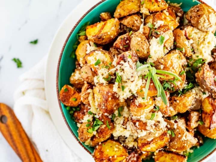 A bowl of roasted baby potatoes topped with herbs and grated cheese on a white tablecloth, with a wooden spoon beside it.