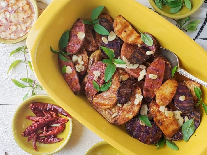 A yellow dish filled with roasted mixed baby potatoes garnished with herbs and garlic slices. Nearby are bowls of chili peppers, sauce, and garlic, making it a perfect addition to your favorite baby potatoes recipes.