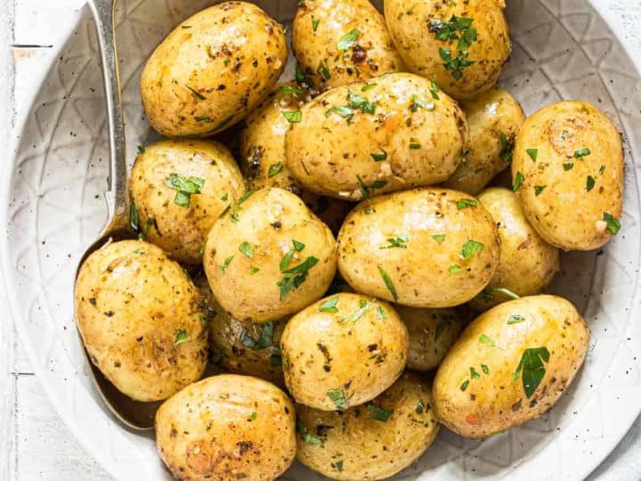 A bowl of roasted baby potatoes, a staple in delicious baby potato recipes, is garnished with herbs and placed on a white wooden surface, with a spoon resting beside it.