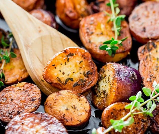 Close-up of roasted baby potato slices with herbs, garnished with sprigs of thyme and stirred with a wooden spoon.