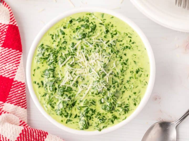 A bowl of creamy spinach soup, an ideal side for lamb chops, is topped with shredded cheese. It's placed invitingly on a white table next to a red and white checkered napkin and a gleaming spoon.