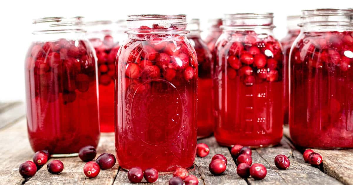 Several glass jars filled with red cranberries and liquid are placed on a wooden surface. Scattered cranberries surround the jars.