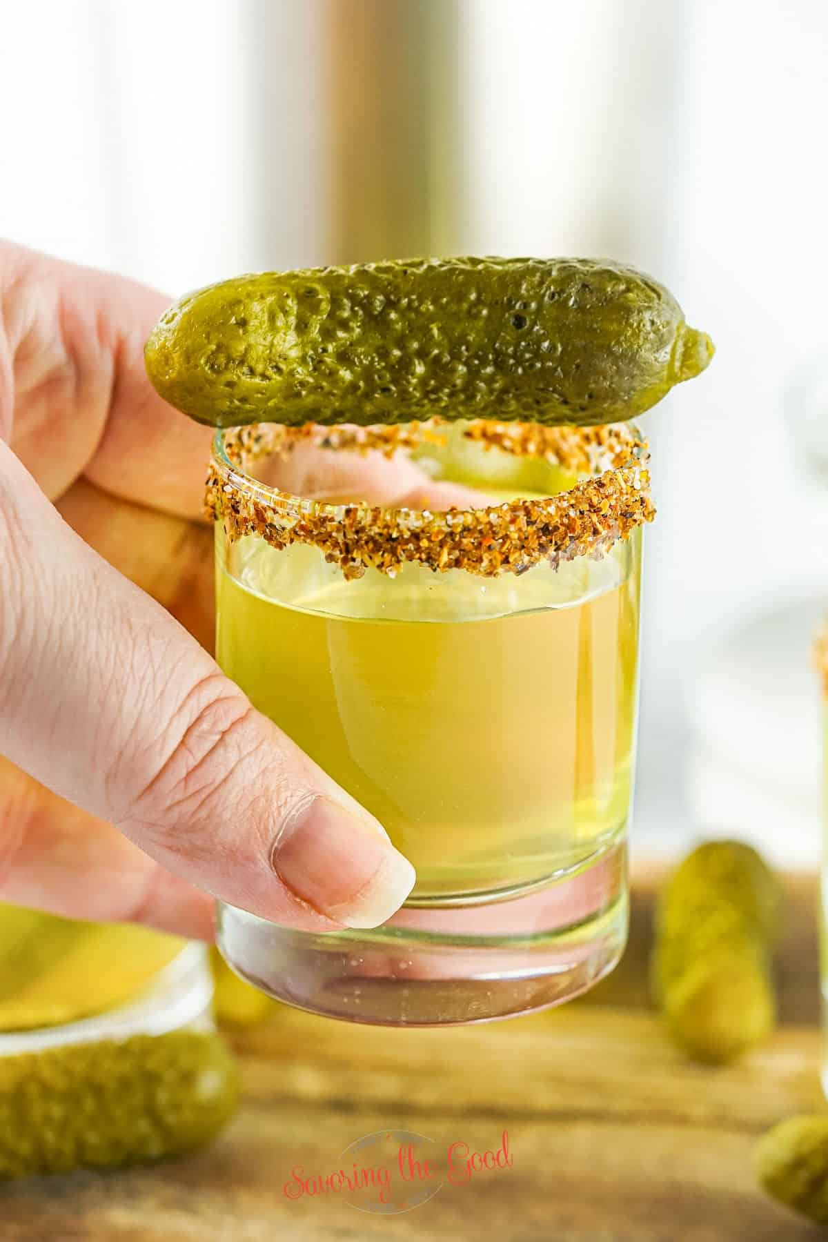 Pickle shot with tajhin rim and gerkin garnish in a clear glass being held by a hand.