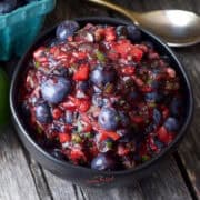 A delightful blueberry salsa recipe awaits you, with a colorful bowl of blueberries and chopped red fruit, elegantly garnished with fresh herbs. All this vibrant goodness sits on a rustic wooden surface, patiently waiting beside a spoon for your enjoyment.