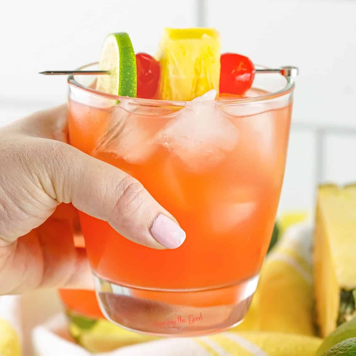 rum punch with garnishes in a clear rocks glass being held by a female hand.