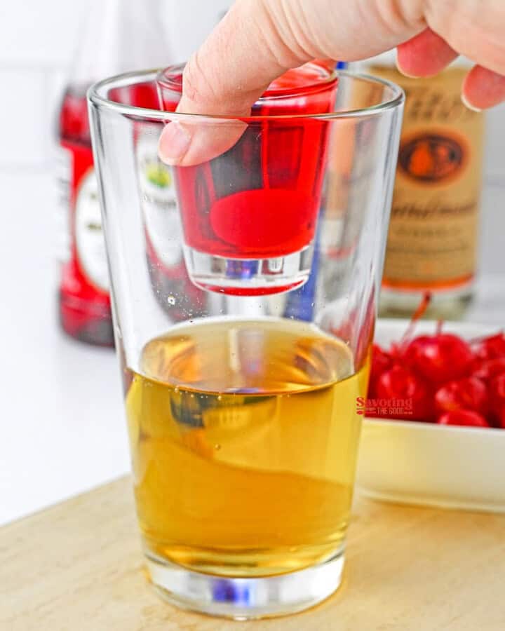 A person's hand dropping a cherry into a glass of amber-colored liquid, with a bottle and bowl of cherries in the background for preparing a cherry bomb shot.