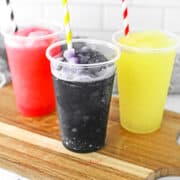 Three plastic cups filled with red, black, and yellow slushie drinks, each with a striped straw, are placed on a wooden board.