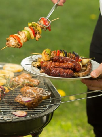 Person holding a plate with grilled skewers and sausages next to a barbecue grill with more skewers cooking outdoors, perfect for Memorial Day recipes.