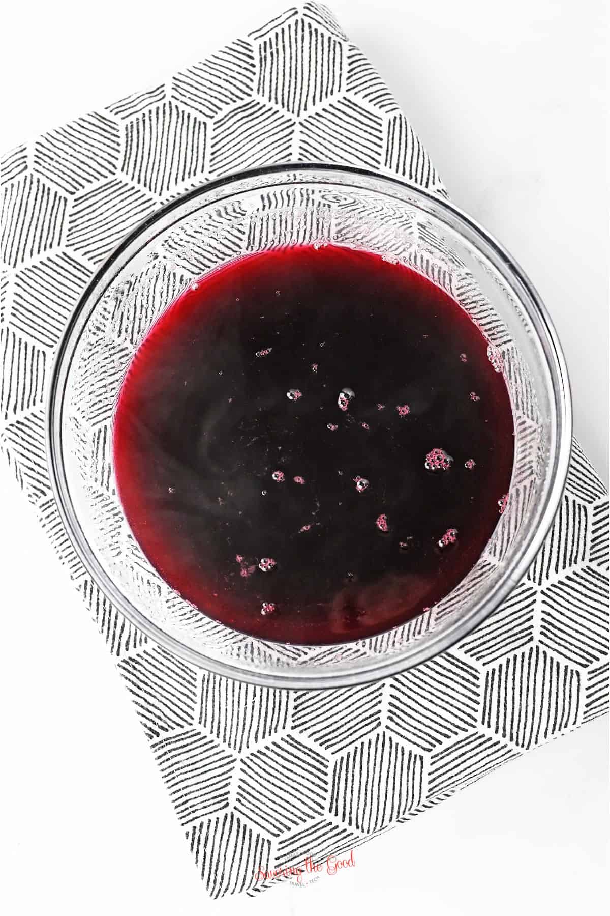 A clear glass bowl filled with dark red liquid sits on a patterned black and white napkin against a white background.