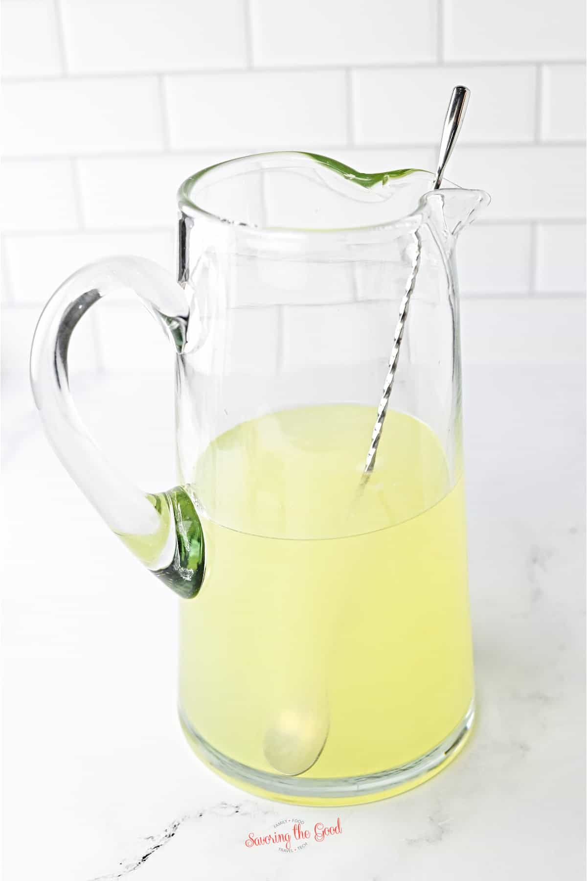 A glass pitcher filled with yellow lemonade and a metal stirring spoon, set on a white surface with a white tiled background.