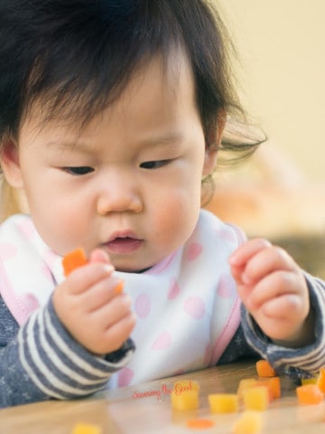 healthy baby snacks featured image of a baby holding tinny carrot pieces.