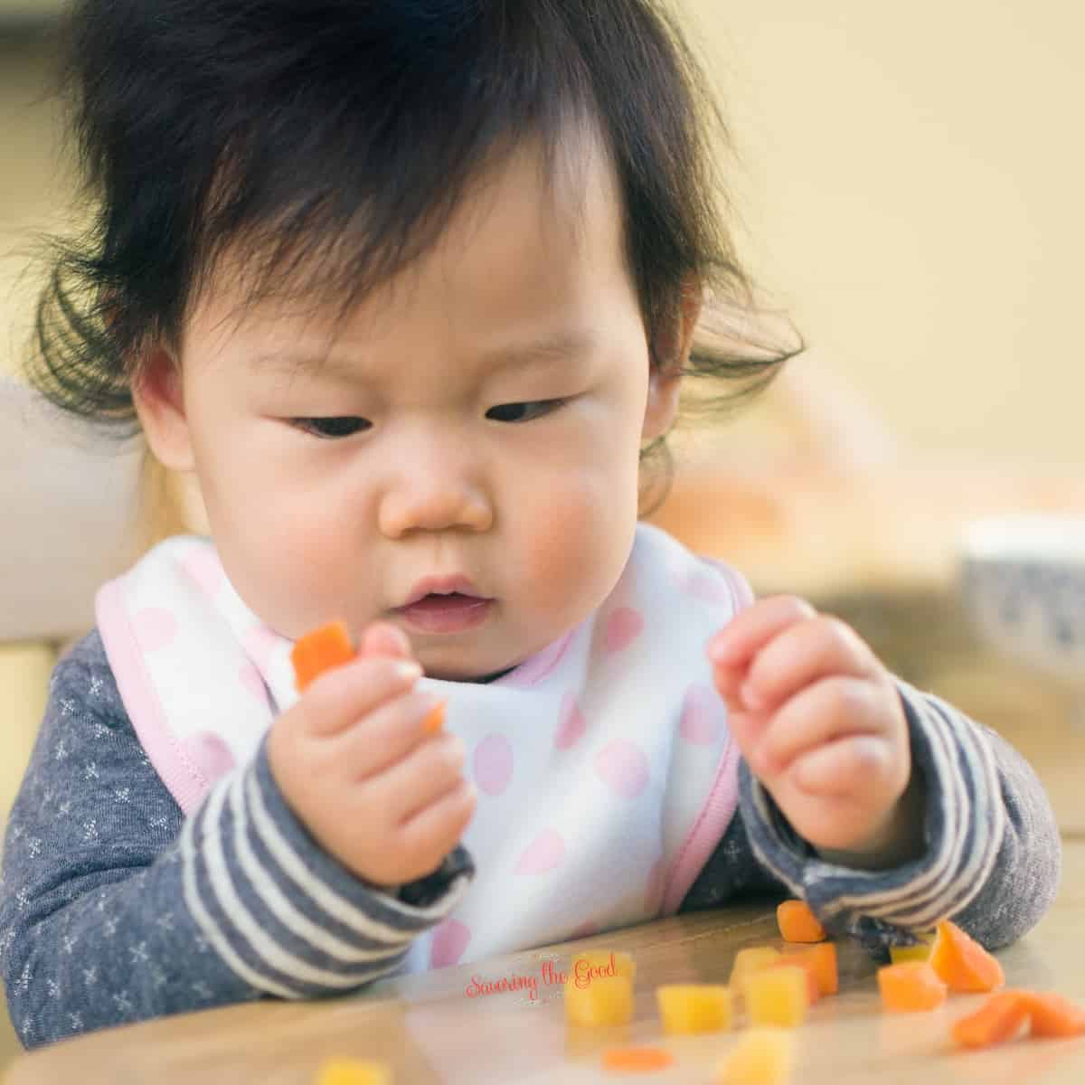 healthy baby snacks featured image of a baby holding tinny carrot pieces.