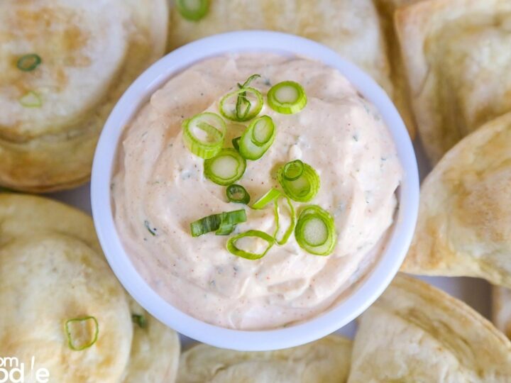 A bowl of creamy sour cream dip topped with sliced green onions, surrounded by baked pierogi on a plate.