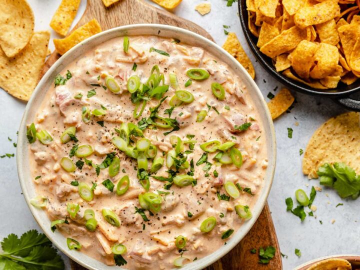 A bowl of creamy sour cream dip topped with chopped green onions and herbs, surrounded by tortilla chips and corn chips on a light surface.