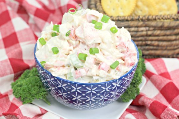 A blue patterned bowl filled with creamy crab salad-reminiscent of classic sour cream dips-topped with chopped green onions, set on a red and white checkered cloth with crackers in the background.