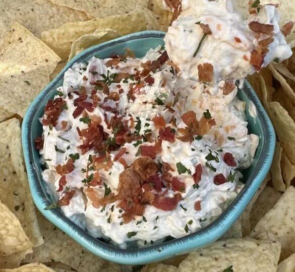 A bowl of creamy sour cream dip topped with bacon bits and herbs, surrounded by tortilla chips, with one chip scooping up the flavorful dip.