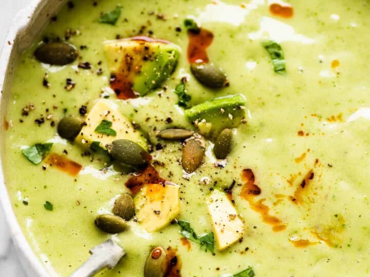 A bowl of green soup garnished with avocado chunks, pumpkin seeds, herbs, and a red seasoning. A spoon lies in the bowl-perfect for those wondering what to serve with sea bass.