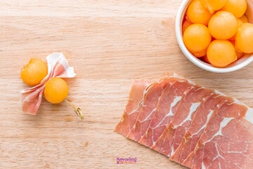 A bowl of melon balls with procuitto on a wooden table.