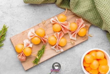 cantaloupe balls wrapped with ham and rosemary on a wooden cutting board.