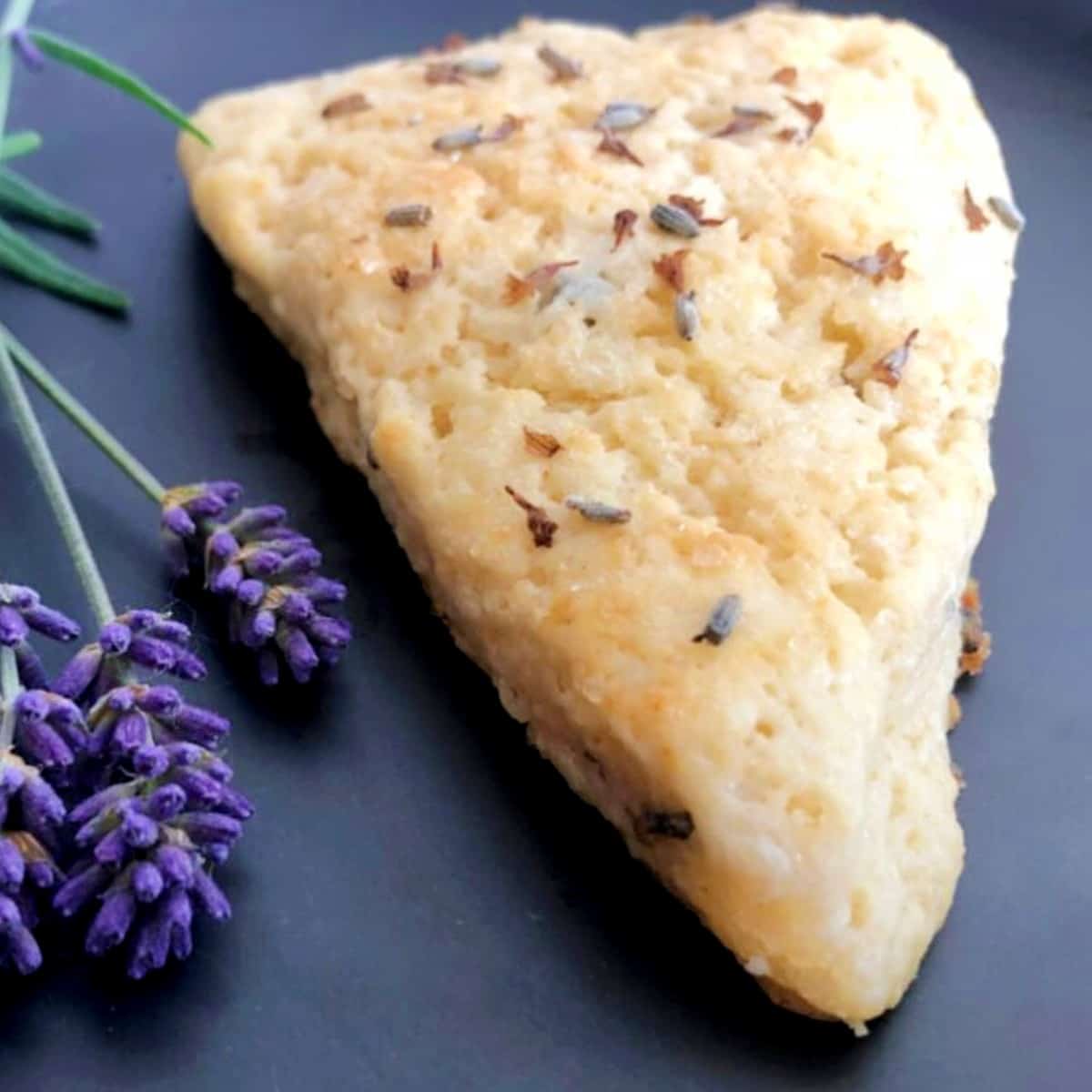 A slice of scone with lavender on a black plate.