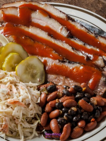 Sliced barbecue pork with coleslaw, beans, pickles, and onion on a plate.
