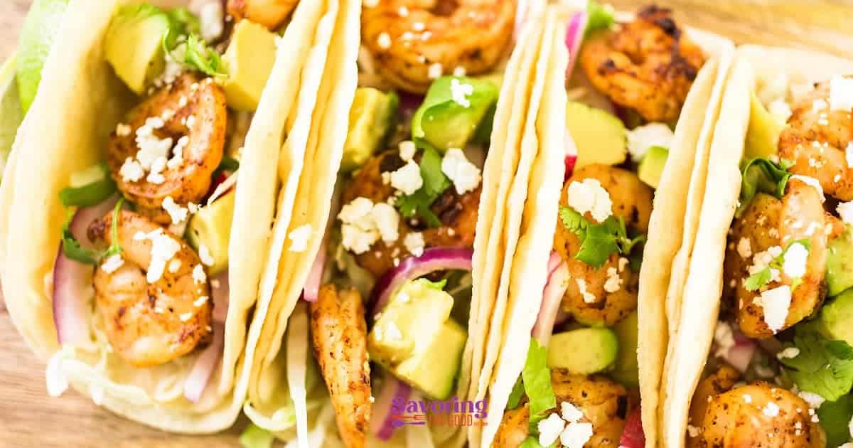 A row of shrimp tacos with avocado and toppings on a wooden surface.