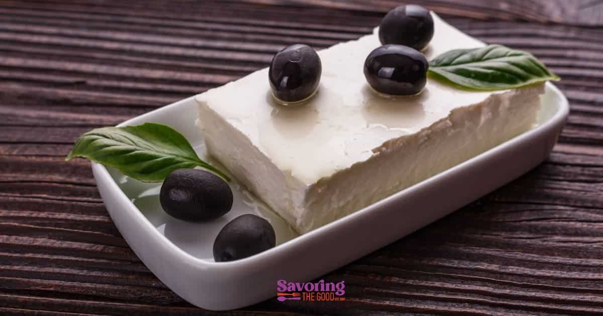 A block of feta cheese garnished with basil leaves and black olives on a small white dish.