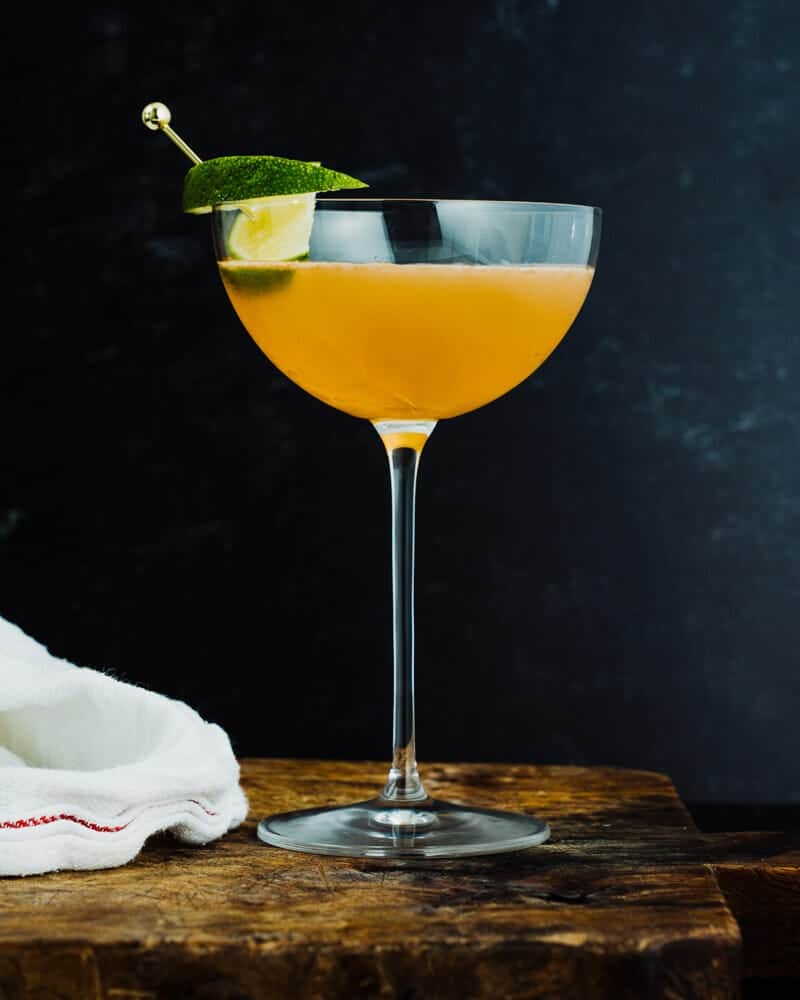 A cocktail with a lime garnish on a wooden cutting board.