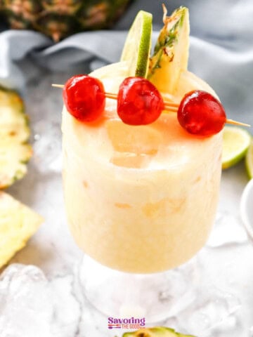 A frosty pineapple mocktail garnished with cherries and a pineapple slice, served on a marble surface surrounded by ice cubes, pineapple chunks, and lime wedges.