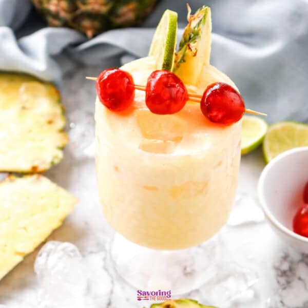 A frosty pineapple mocktail garnished with cherries and a pineapple slice, served on a marble surface surrounded by ice cubes, pineapple chunks, and lime wedges.