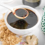 A spoon drizzling Teriyaki Sauce into a small bowl, with ginger and a jar of soy sauce in the background.