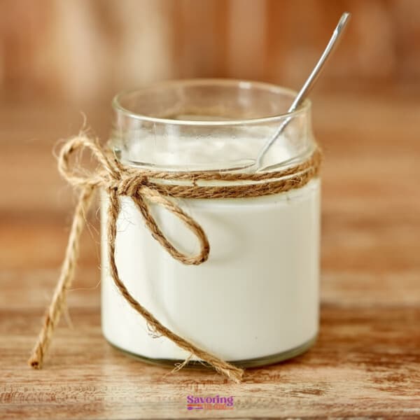 Clear glass jar filled with double cream, tied with a jute string, with a spoon inside, on a wooden table.
