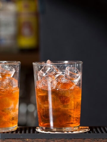 Pouring a brown liquid into two glasses with ice on a bar counter during the 1920 Prohibition era, with a third glass containing a red drink.