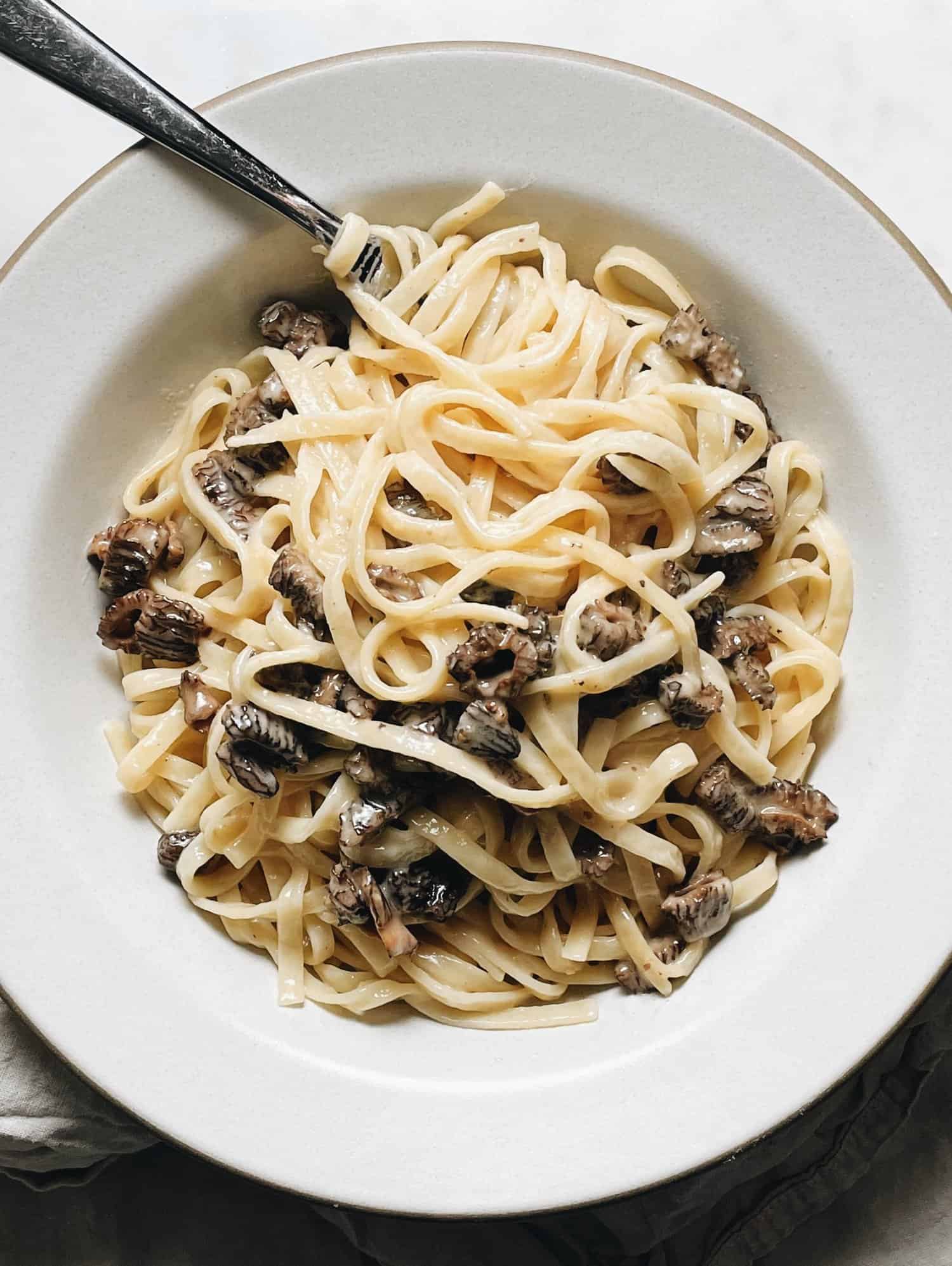 A white bowl of fettuccine pasta with a creamy sauce and mushrooms, with a fork resting in the bowl.
