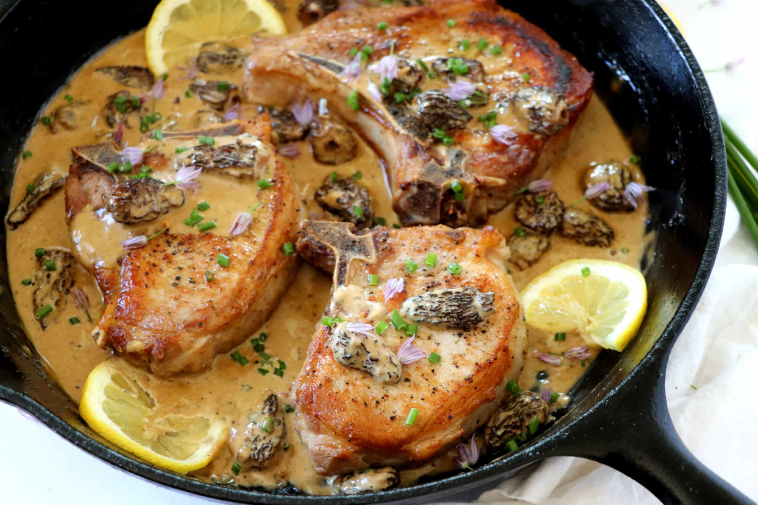 Three seared pork chops in a creamy sauce with morel mushrooms, lemon slices, and chopped herbs, served in a black cast iron skillet.
