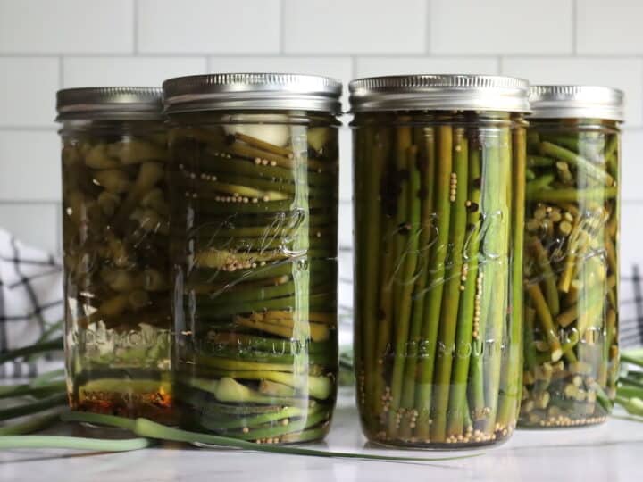 Four glass jars filled with pickled green scapes are sealed with metal lids, lined up on a counter with a checkered cloth and white subway tile background.