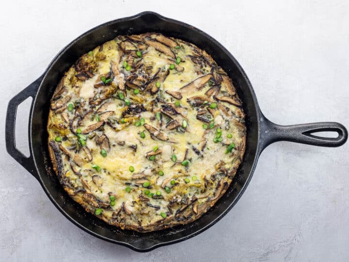 A baked frittata with mushrooms, cheese, green peas, and tender scapes in a black cast iron skillet on a light-colored surface.