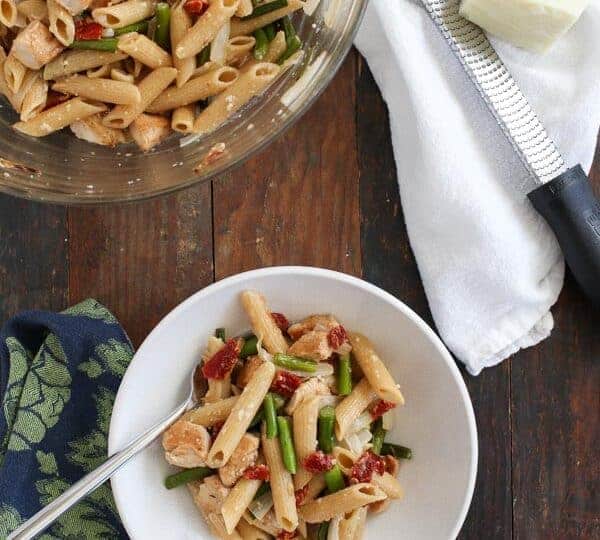 A bowl and a large glass bowl of penne pasta with chicken, green beans, scapes, and sun-dried tomatoes sit on a wooden table, next to a grater, cheese, and a white towel.