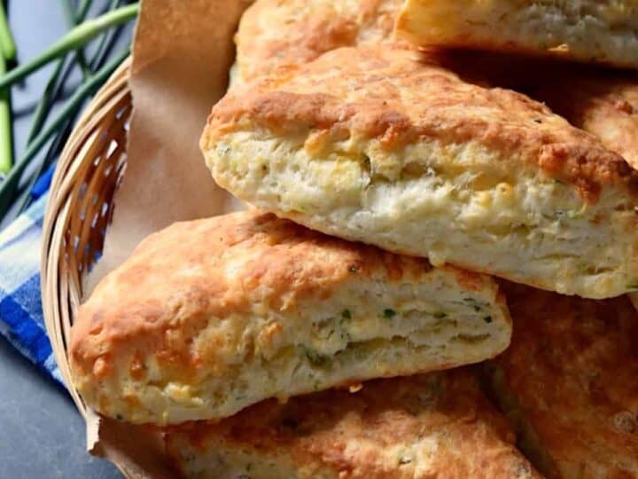 A basket filled with several golden-brown, triangular cheese scones stacked on top of each other, flecked with tender green scapes for a fresh twist.